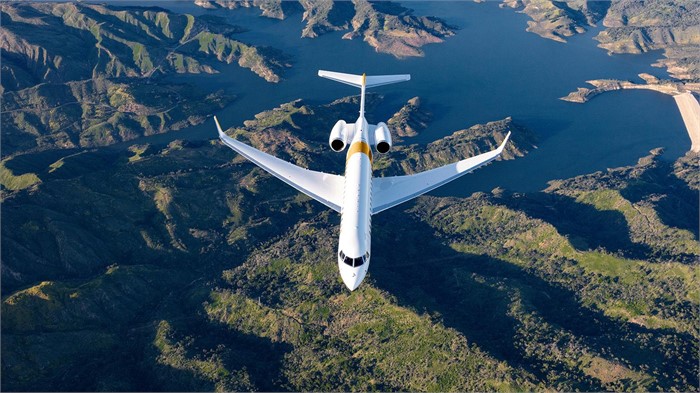A white Bombardier Global 7500 business jet shown from above, flying over water and rugged hills.