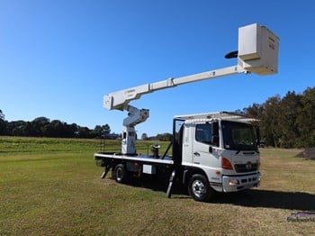 A white Hino cherry picker truck with jack supports for stabilization and a basket or work platform extended on an elevating boom.