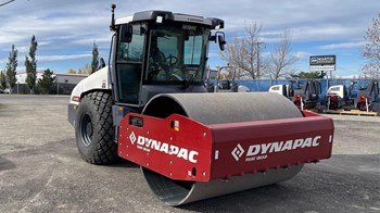 A white and red Dynapac smooth-drum compactor sitting in an equipment lot.