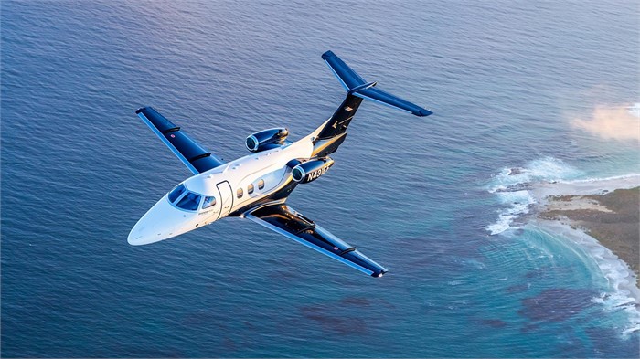 A twin-engine Embraer Phenom 100EX light jet flies along an ocean coastline with clouds in the background.