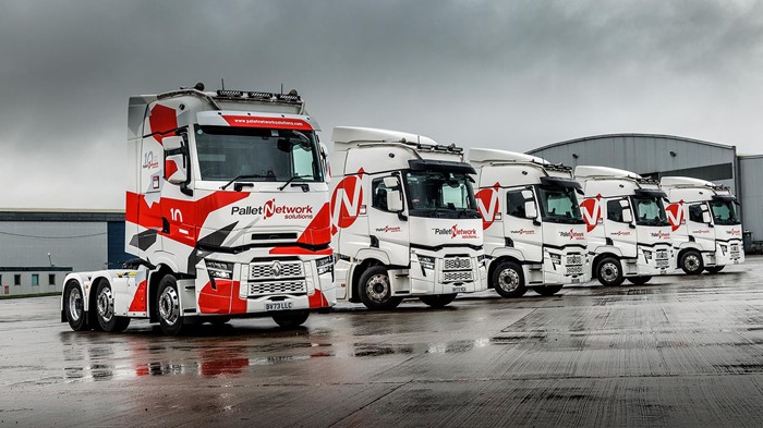 Five of Pallet Network Solutions’ white-and-red Renault tractor units sitting in a row on a large, wet, paved surface.